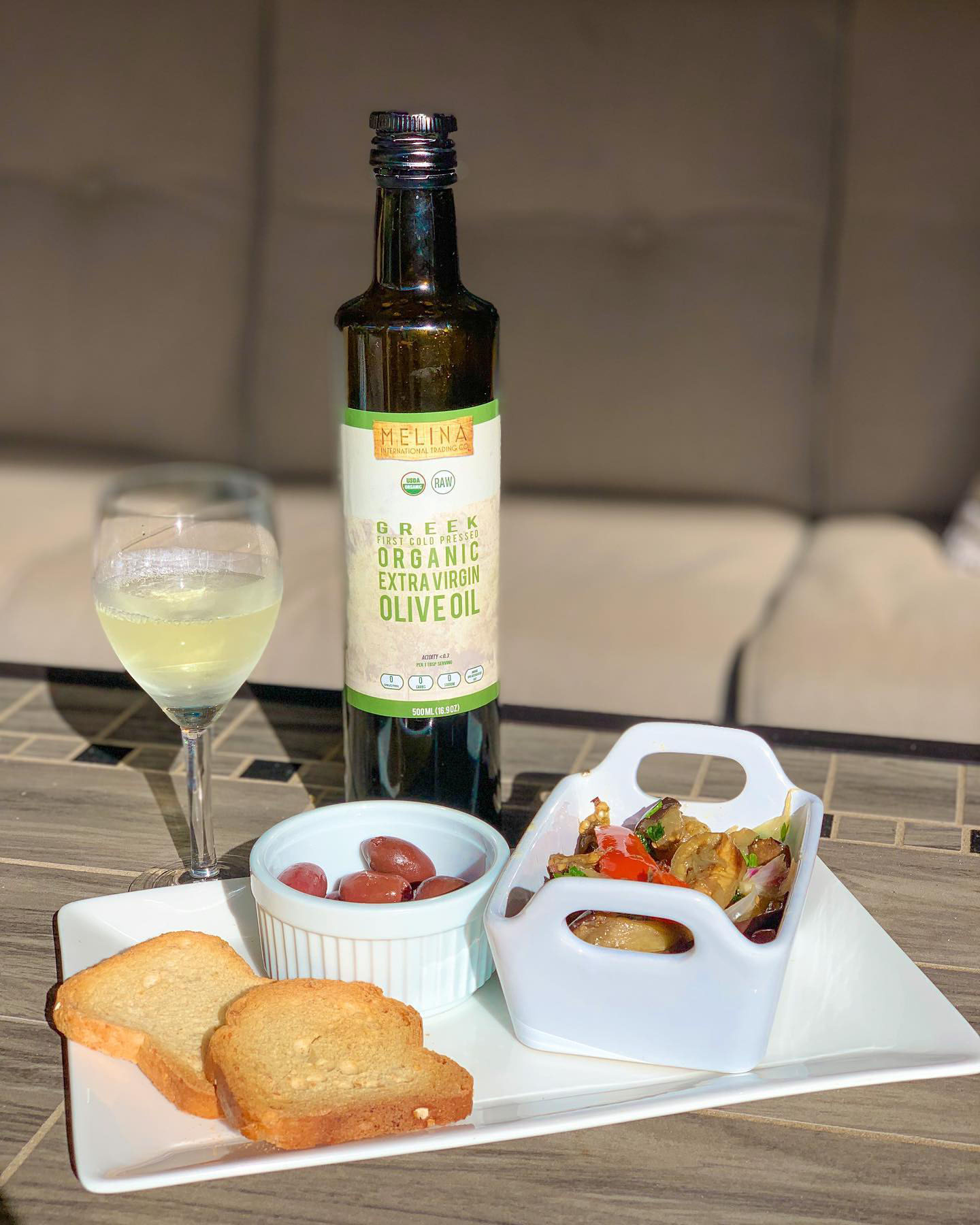 Bottle of Melina Extra Virgin Olive Oil next to olives, salad, and wine